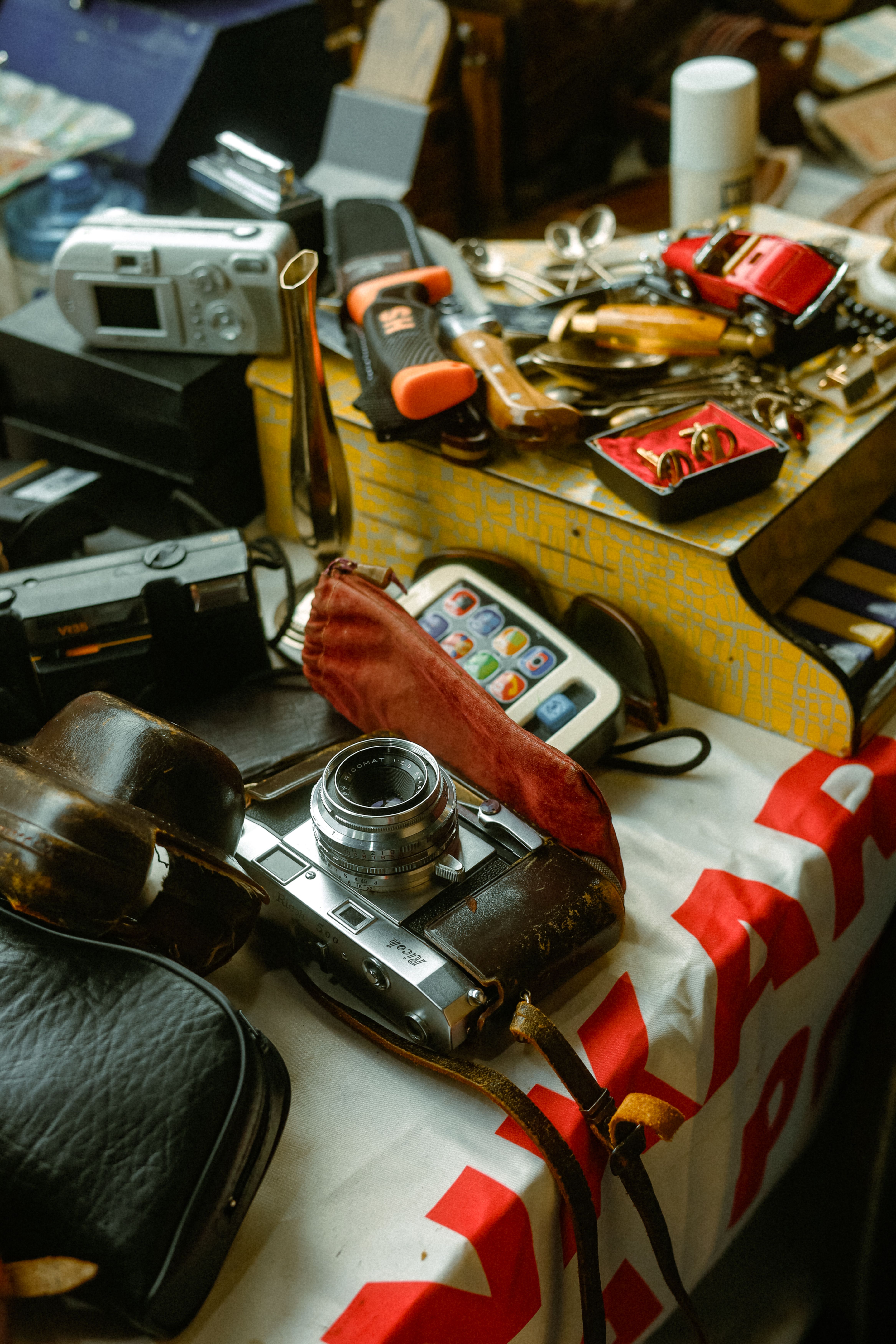 Flea market table with a vintage Ricoh rangefinder camera in a worn leather case, older digital cameras, cufflinks, spoons and a red toy car