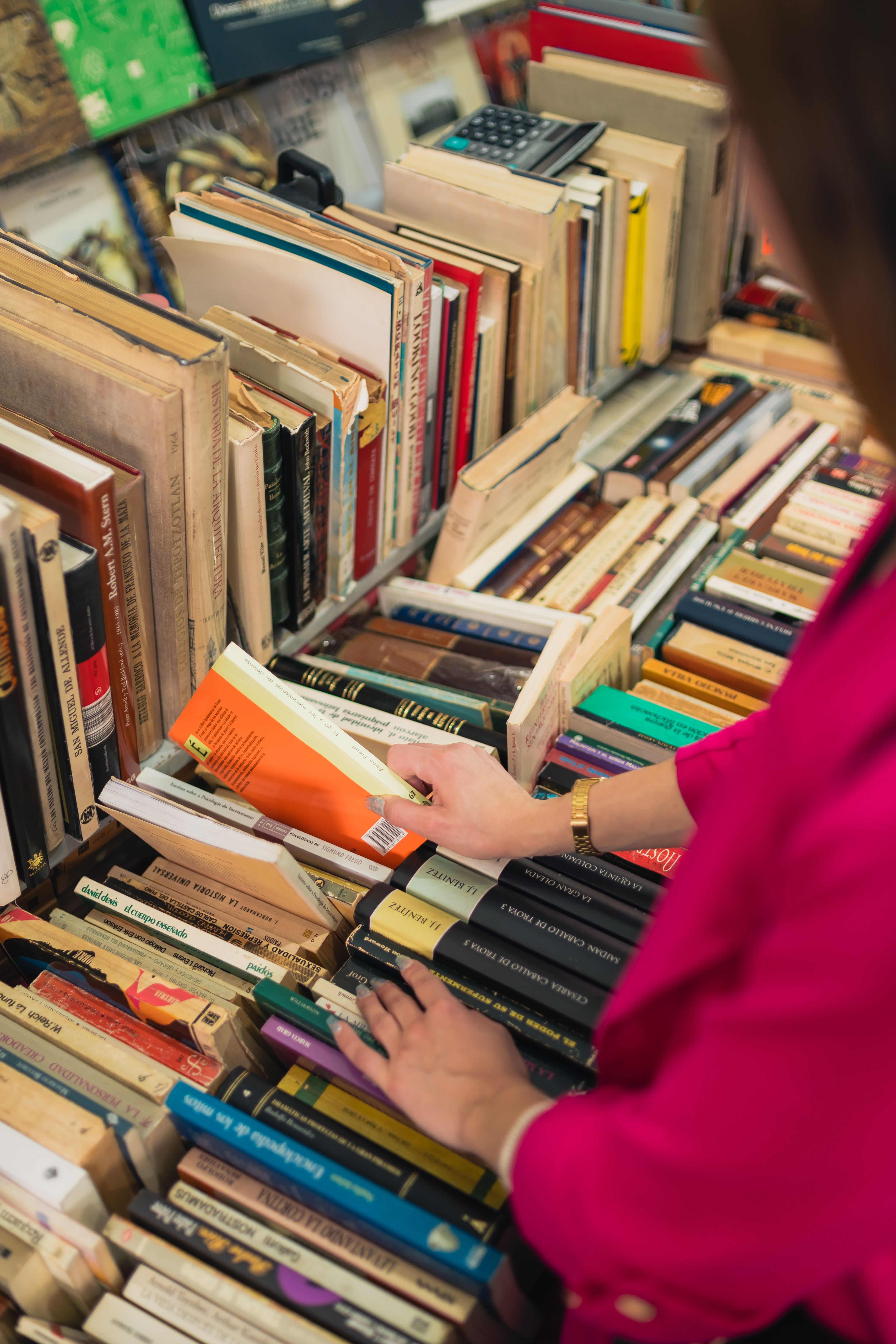A shopper in a pink sweater pulls an orange paperback from a crowded used-book table, with stacks of hardcovers and a calculator on the shelf behind