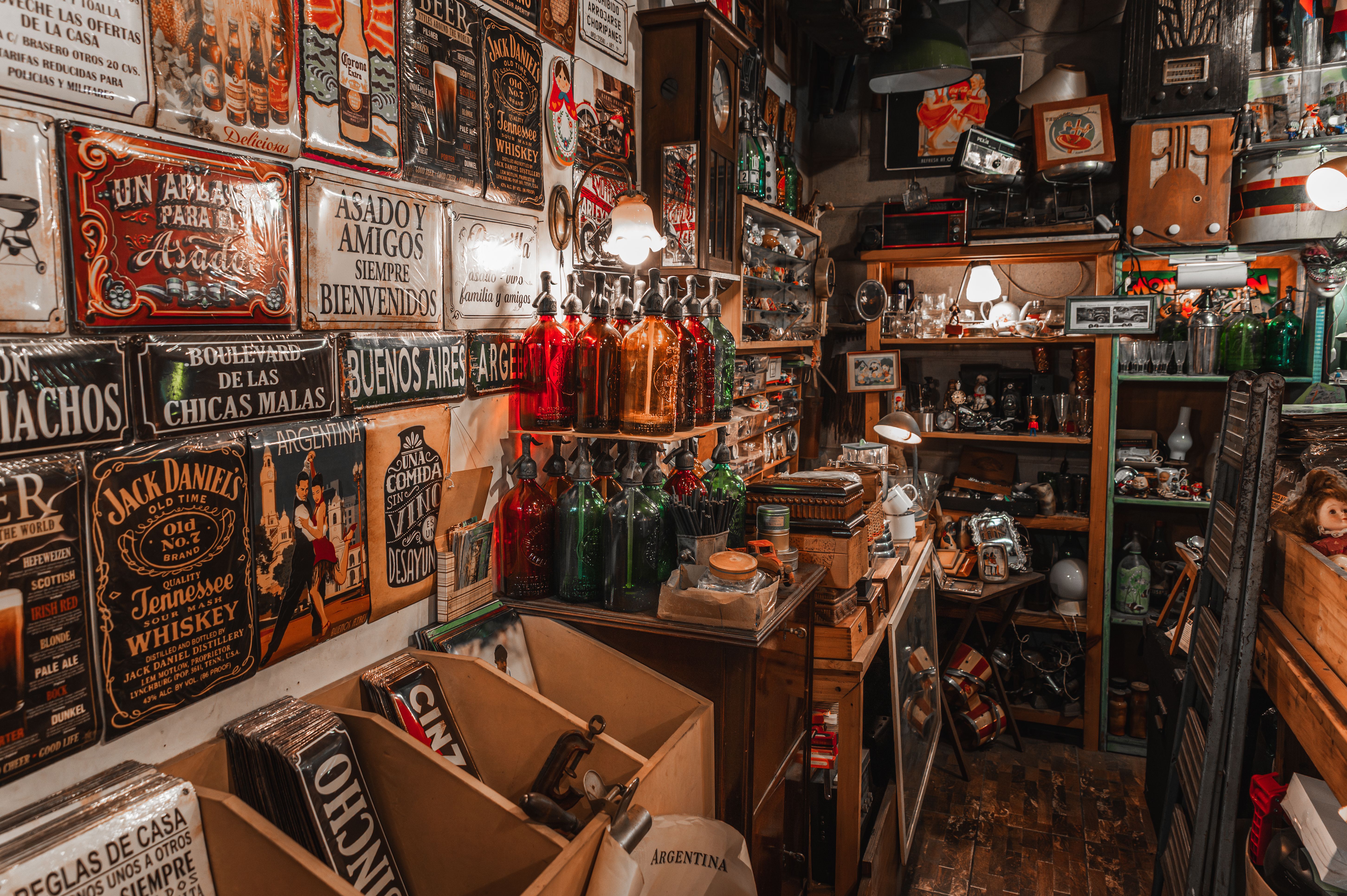 Crowded vintage shop with tin signs on the wall, colored glass seltzer bottles, shelves of radios, clocks and collectibles, and bins of stock