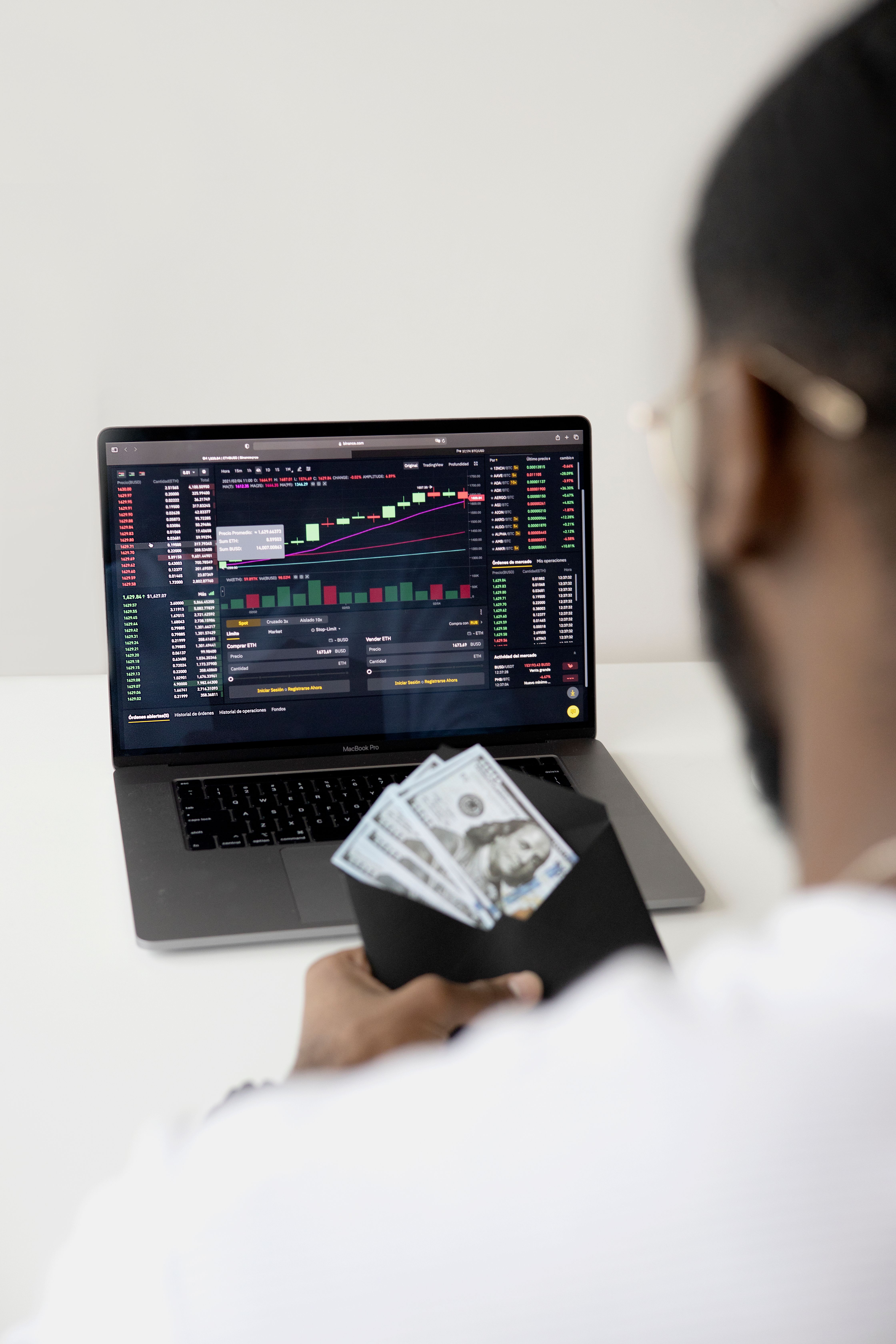 Person at a desk holding a black envelope of US hundred-dollar bills while looking at a laptop showing a price chart