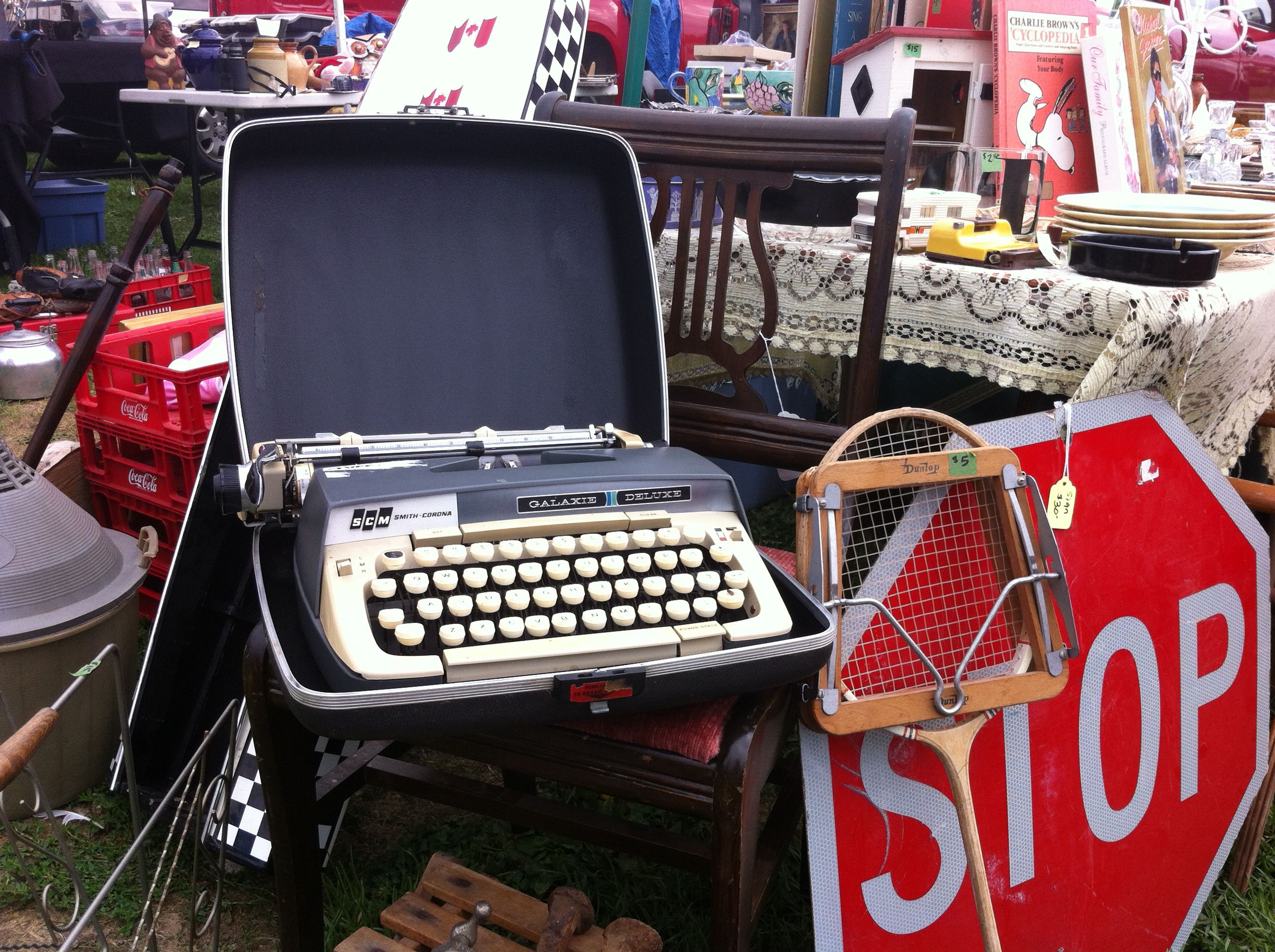 A vintage portable typewriter in an open case on a wooden chair at an outdoor flea market, with a stop sign, a wooden tennis racket, and tables of secondhand goods behind it