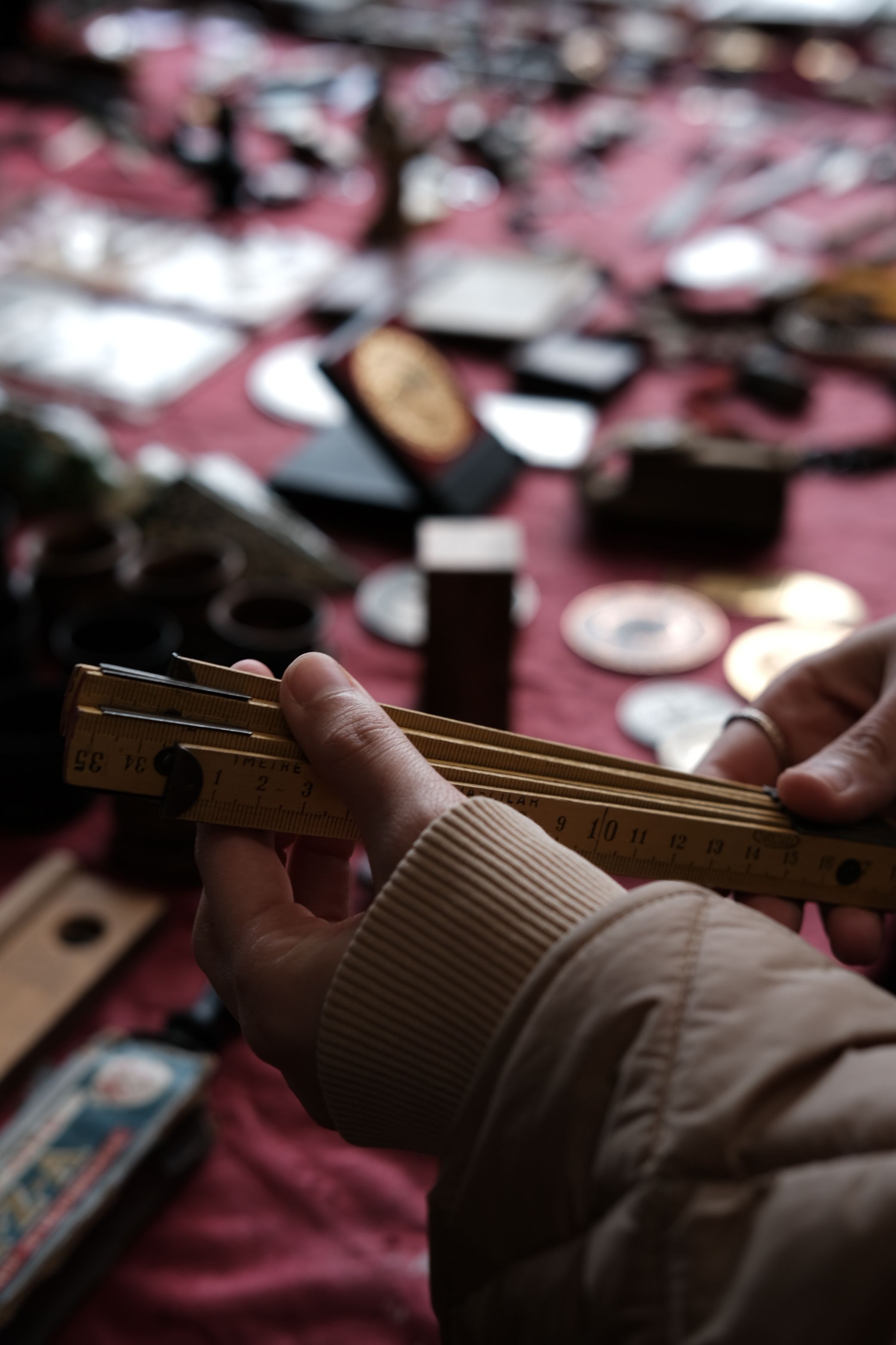 Hands holding a vintage wooden folding ruler over a flea market table covered in coins, medals and small collectibles